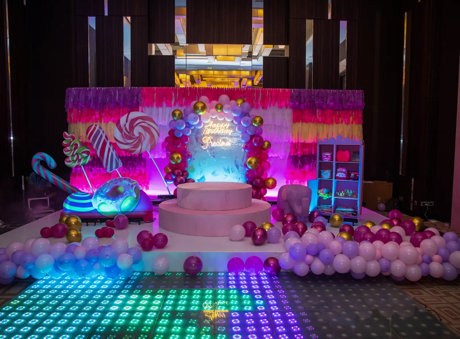 Candyland themed birthday stage with giant lollipops, a neon birthday sign, a pink and gold balloon arch and an LED dance floor