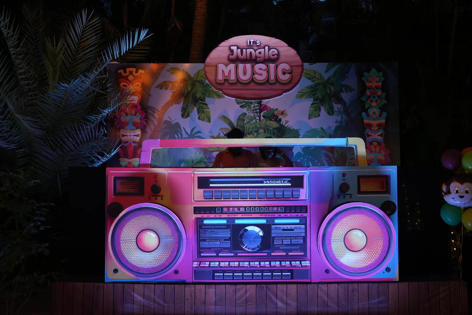 Outdoor jungle party DJ booth built as an oversized retro boombox, lit in pink and blue against a tropical backdrop