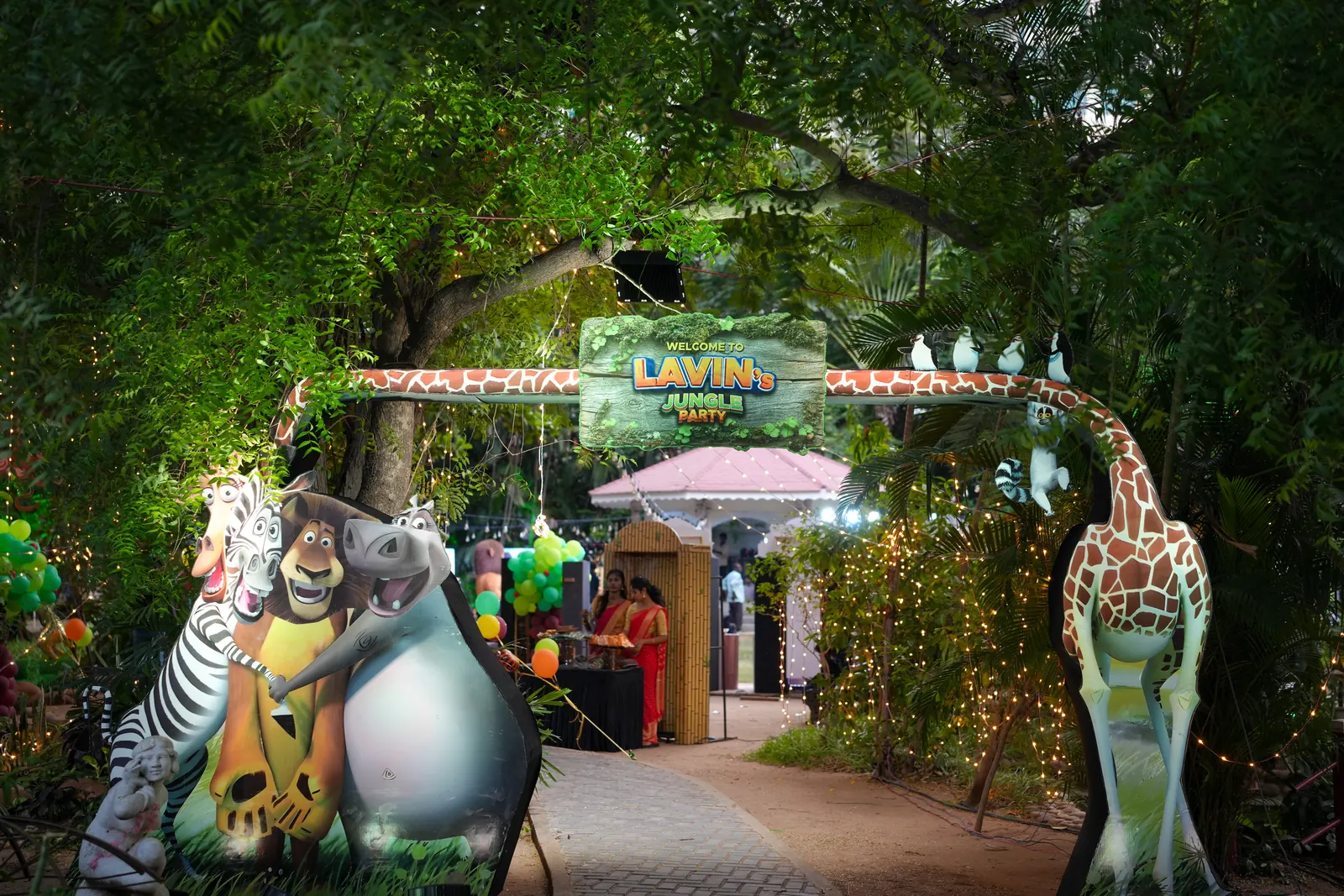 Garden pathway into a jungle themed party at dusk, lined with animal cut-outs, a giraffe arch, fairy lights and balloon clusters
